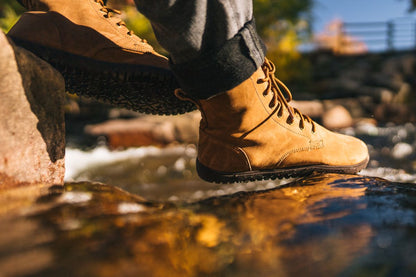 Person wearing brown boots standing on a rock by a stream.  Tru Stride Co brings Xero Shoes to Canada