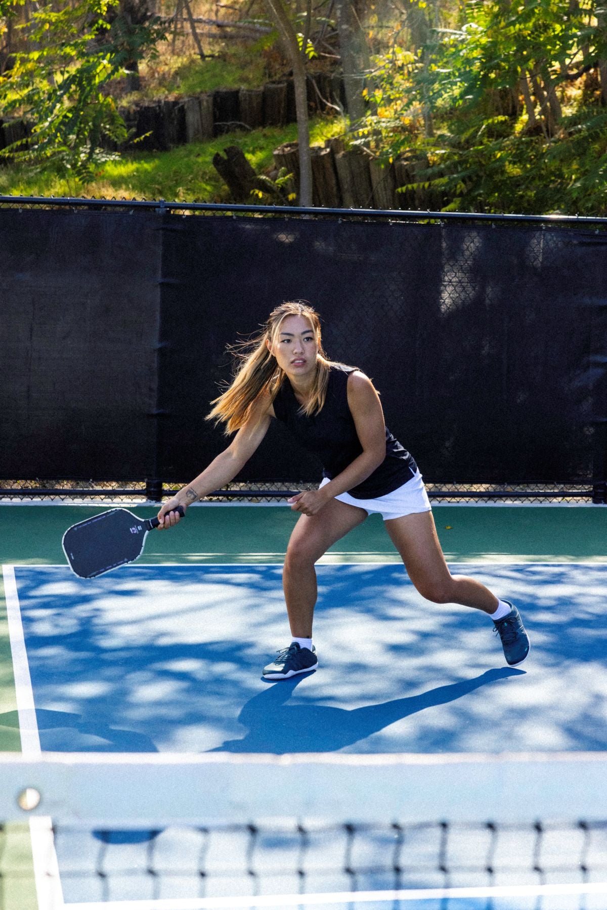 Woman playing pickleball on an outdoor court with trees in the background.  Tru Stride Co brings Xero shoes to Canada